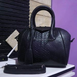 Könnte beinhalten: Eine schwarze Lederhandtasche mit einem strukturierten Schlangenmuster. Die Tasche hat einen oberen Griff, einen abnehmbaren Schultergurt und eine Quaste. Die Tasche steht auf einer weißen Oberfläche.