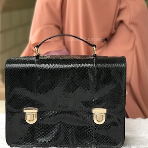 Könnte beinhalten: Schwarze Lederhandtasche mit Schlangenmuster, einem oberen Griff und zwei goldfarbenen Verschlüssen. Die Tasche hat eine rechteckige Form und ein strukturiertes Design. Der Text "SUMI LEATHER" ist oben auf der Tasche zu sehen.