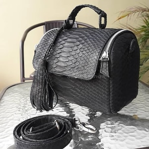 Könnte beinhalten: Schwarze Lederhandtasche mit strukturierter Schlangenmusterung. Die Tasche hat einen oberen Griff, einen abnehmbaren Schulterriemen und eine Quaste. Der Reißverschluss und die Beschläge sind silberfarben. Auf einem Glastisch.