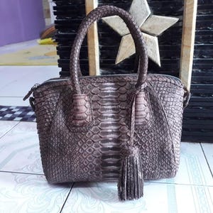 Könnte beinhalten: Eine braune Handtasche aus Schlangenleder mit einem oberen Griff und einem Reißverschluss. Die Tasche hat eine Quaste und ein strukturiertes Muster. Die Tasche steht auf einer gefliesten Oberfläche.