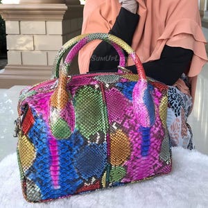 May include: A vibrant handbag with a snakeskin design. The bag displays a mix of colours, such as pink, blue, green, and yellow. It has two handles and a zip closure. The text "SUMUPL" is visible on the handle.