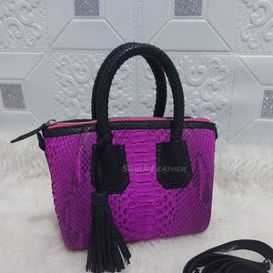 May include: A bright purple handbag with a snakeskin texture, black handles, and a detachable shoulder strap. The bag has a black tassel and a pink zip. The brand name "SUMUDLEATHER" is visible on the front.