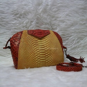 Könnte beinhalten: Eine rote, gelbe und beige Handtasche mit Schlangenmuster, passendem Riemen und Quaste. Die Tasche hat eine abgerundete Form mit einem Klappenverschluss und einem abnehmbaren Riemen. Die Tasche ist aus einem strukturierten Material gefertigt.