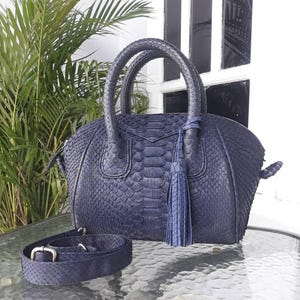 Könnte beinhalten: Eine marineblaue Handtasche mit einer strukturierten, schlangenlederartigen Musterung. Die Tasche hat zwei runde Griffe, einen abnehmbaren Schulterriemen und eine Quaste. Die Tasche steht auf einem Glastisch.