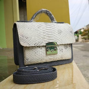 Könnte beinhalten: Eine Handtasche mit weißem und grauem Schlangenmuster, einem silbernen Verschluss und einem grauen Riemen. Die Tasche hat einen oberen Griff und einen Klappenverschluss. Die Seiten und der Boden der Tasche sind grau.