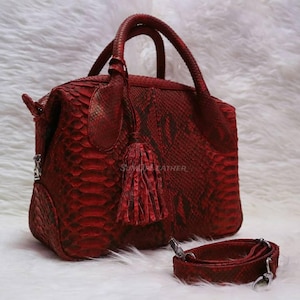 Könnte beinhalten: Eine rote Lederhandtasche mit Schlangenmuster, mit einem oberen Griff, einem abnehmbaren Schulterriemen und einer dekorativen Quaste. Die Tasche hat silberfarbene Hardware und einen Reißverschluss. Die Tasche ist etwa 30 cm breit.