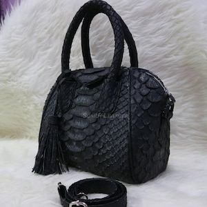 Könnte beinhalten: Schwarze Lederhandtasche mit einem strukturierten, schuppenartigen Muster. Die Tasche hat zwei Griffe, einen Reißverschluss und einen abnehmbaren Schulterriemen. Eine Quaste hängt an der Seite. Die Tasche steht auf einer weißen, flauschigen Oberfläche.