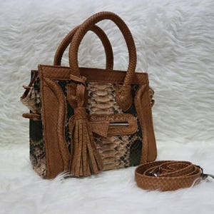 Könnte beinhalten: Eine braune und schwarze Handtasche mit Schlangenmuster, mit einem oberen Griff und einem abnehmbaren Schultergurt. Die Tasche hat eine Fronttasche mit Reißverschluss, eine Quaste und den Text "SUMU LEATHER".