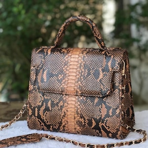 Op de afbeelding: Een bruine en zwarte handtas van slangenleer met een handvat aan de bovenkant en een gouden kettingriem. De tas heeft een rechthoekige vorm met een klepsluiting en een textuurpatroon. De tas staat op een wit oppervlak.