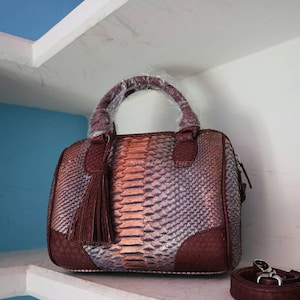 Könnte beinhalten: Handtasche mit Schlangenmuster in Burgunder-, Silber- und Pfirsichtönen. Die Tasche hat einen oberen Griff, eine Quaste und einen abnehmbaren Schultergurt. Die Tasche ist etwa 30 cm breit.