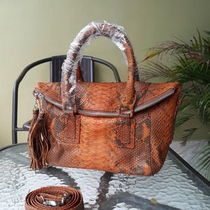 Op de afbeelding: Een oranje en bruine handtas van slangenleer met een handvat aan de bovenkant en een afneembare schouderriem. De tas heeft een ritssluiting en een kwastje. De riem is opgerold en heeft zilverkleurige hardware.
