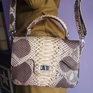 Könnte beinhalten: Rechteckige Handtasche mit Schlangenmuster in Braun- und Beigetönen. Die Tasche hat einen oberen Griff und einen langen, verstellbaren Riemen. Der Verschluss ist silberfarben. Der Text "SUMUPLEATHER" ist auf der Tasche zu sehen.