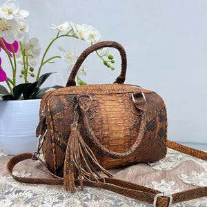 Könnte beinhalten: Braune und hellbraune Handtasche aus Schlangenleder mit einem oberen Griff und einem abnehmbaren Schulterriemen. Die Tasche hat eine Quaste und silberfarbene Hardware. Die Tasche liegt auf einer Spitzendecke.