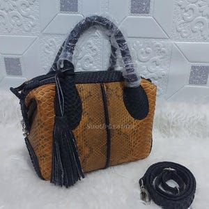 May include: A brown and black leather handbag with a top handle and a detachable shoulder strap. The bag features a tassel and the text "SUNUP LEATHER". The bag is made of a snakeskin pattern.
