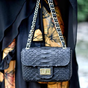 Könnte beinhalten: Eine schwarze Handtasche aus strukturiertem Leder mit Klappenverschluss und goldfarbenem Verschluss. Die Tasche hat einen Kettenriemen mit schwarzen Lederakzenten. Der Hintergrund zeigt ein schwarzes Kleidungsstück und gemusterten Stoff.