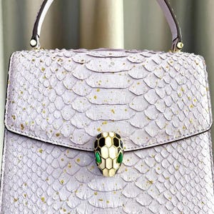 May include: A lilac handbag with a scale-like texture and gold specks. The bag has a top handle and a snake head clasp with black, white, and green details. The bag is held by a person.