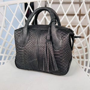 May include: A black leather handbag with a snakeskin texture. The bag features two rounded handles and a tassel. The bag is placed on a white chair.