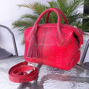 Könnte beinhalten: Eine rote Handtasche mit Schlangenmuster, mit einem oberen Griff und einem abnehmbaren Schulterriemen. Die Tasche hat eine Quaste und einen Reißverschluss. Die Tasche steht auf einem Glastisch.