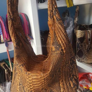 Könnte beinhalten: Eine große, braune Lederhandtasche mit einem markanten Schlangenhautmuster. Die Tasche hat eine geschwungene Form, einen Reißverschluss und einen Schulterriemen. Die Tasche wird in einem Geschäft ausgestellt, mit anderen Lederwaren im Hintergrund.