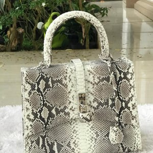 Könnte beinhalten: Rechteckige Handtasche mit Schlangenmuster in Grau-, Weiß- und Schwarztönen. Die Tasche hat einen weißen Griff, eine weiße Klappe mit silbernem Verschluss und einen passenden Riemen mit kleinem Anhänger. Die Tasche liegt auf einer weißen Oberfläche.