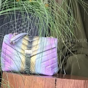 May include: A colorful, quilted handbag with a chain strap. The bag features a mix of purple, blue, and yellow hues, with a central panel of darker, textured material. A gold-toned clasp secures the flap.