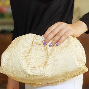Könnte beinhalten: Eine cremefarbene Clutch mit einem strukturierten, schlangenlederartigen Muster. Die Tasche wird von einer Person gehalten, die ein schwarzes Langarmshirt und eine weiße Hose trägt. Die Clutch ist der Blickfang und zeigt ihr einzigartiges Design.