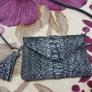 Könnte beinhalten: Dunkelgraue Clutch mit Schlangenmuster, geflochtenem Riemen und Quaste. Die Tasche hat einen Klappverschluss und einen silberfarbenen Verschluss. Der Hintergrund zeigt ein Blumenmuster in Lila und Beige.