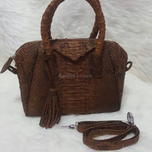 Könnte beinhalten: Braune Lederhandtasche mit strukturierter Schlangenmusterung. Die Tasche hat einen oberen Griff, einen abnehmbaren Schulterriemen und eine Quaste. Der Text "SUMUPLEATHER" ist auf der Vorderseite der Tasche sichtbar.