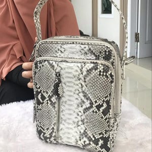 Könnte beinhalten: Quadratische Umhängetasche mit Schlangenmuster in Grau- und Weißtönen. Sie hat eine Vordertasche mit Reißverschluss, einen oberen Reißverschluss und einen Griff. Die Tasche ist aus einem strukturierten Material gefertigt.