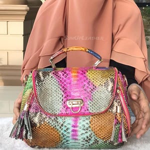 May include: A vibrant handbag featuring a snakeskin design in pink, green, yellow, and brown. It has a top handle, a metal clasp, and a zip with a tassel. The text "SUMUPLEATHER" is visible on the bag.