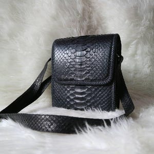 Op de afbeelding: Een zwarte crossbody tas met een rechthoekige vorm en een klep. De tas is gemaakt van een textuur die op slangenleer lijkt en heeft een lange, verstelbare riem. Op een witte, donzige achtergrond.