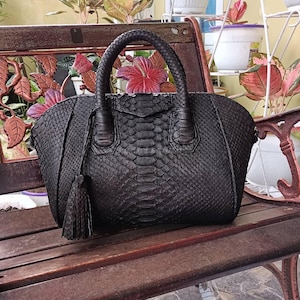 May include: Black handbag with a snakeskin texture, a top handle, and a tassel. The bag is on a wooden bench with floral details. The bag has a structured shape and a flap closure. The bag is made of leather.