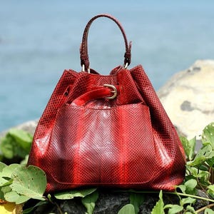 Peut inclure: Un sac à main rouge texturé imitation peau de serpent, avec une poignée supérieure et une fermeture à cordon. Le sac a une poche avant et une boucle dorée. Le sac est posé sur un rocher avec du feuillage vert, devant un océan bleu.