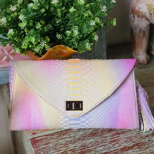 May include: A pastel-coloured clutch bag with a snakeskin pattern. The bag has a flap closure with a rectangular clasp and a tassel on the side. The colour palette includes pink, yellow, and purple hues.