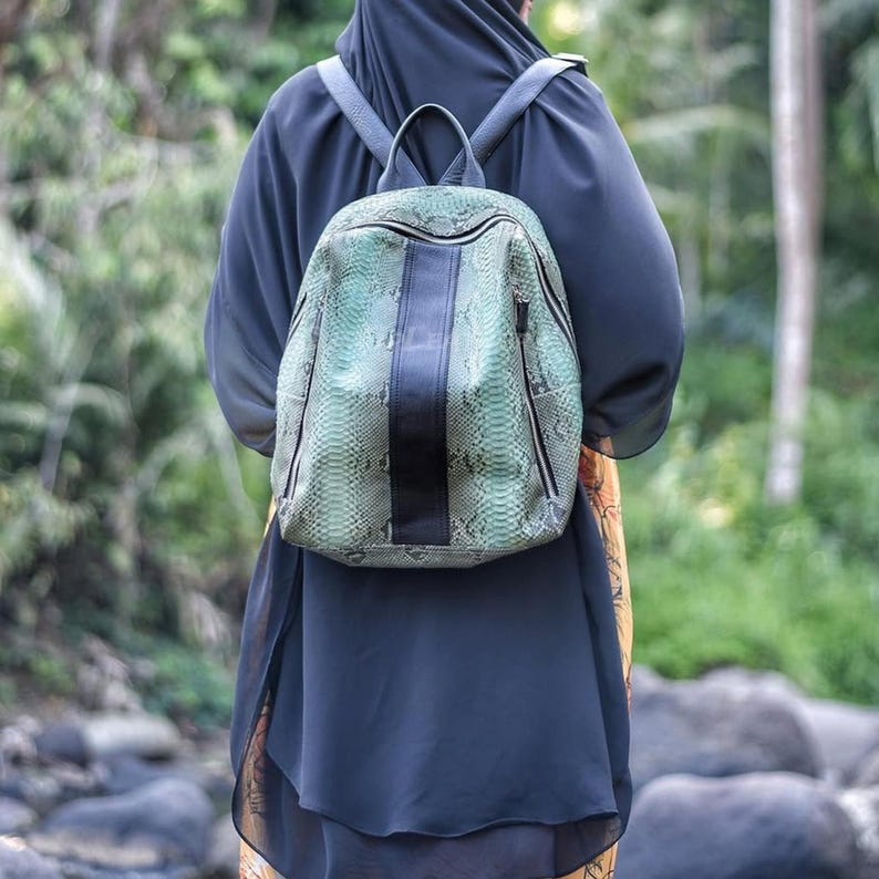 May include: A backpack featuring a textured green and grey snakeskin pattern with a solid black vertical stripe down the center. The bag includes a top handle, two shoulder straps, and two side zippers with dark hardware, worn on the back.