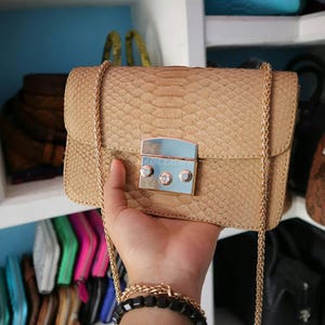May include: A tan, rectangular handbag with a chain strap and a silver clasp. The bag has a snakeskin pattern and is held by a person. The clasp has the text "JP LEATHER" on it. The bag is displayed in a store setting.