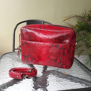 Könnte beinhalten: Eine rote und schwarze Handtasche mit Schlangenmuster und passendem Schulterriemen. Die rechteckige Tasche hat einen Reißverschluss und eine Vordertasche. Der Riemen ist verstellbar und hat Metallbeschläge. Die Tasche steht auf einem Glastisch.