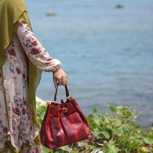 May include: A red leather handbag with a drawstring closure and a textured snakeskin pattern. The bag has a small pocket on the front. The text "SumUpLeather" is visible. The bag is held near the sea.