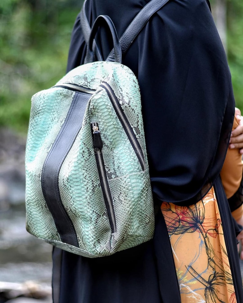 May include: A backpack featuring a mint green and black snakeskin pattern with a vertical black leather-like center stripe. The bag includes black zippers, a top handle, and adjustable shoulder straps, showcasing a textured reptilian finish.