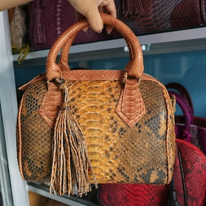 Könnte beinhalten: Handtasche aus braun-gelbem Schlangenhautleder mit oberem Griff und Quaste. Die Tasche hat eine runde Form und einen Reißverschluss. Der Griff und die Verzierungen sind hellbraun.