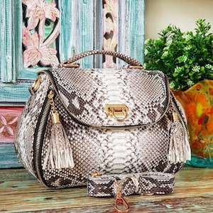 May include: A stylish handbag with a snakeskin pattern in shades of white, gray, and black. The bag features a top handle, a flap closure with a gold-tone clasp, and a detachable shoulder strap. Decorative tassels hang from the zippers.
