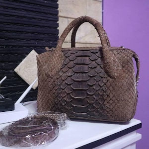 Könnte beinhalten: Eine braune Handtasche aus Schlangenhaut mit zwei Griffen und einem abnehmbaren Schulterriemen. Die Tasche hat eine rechteckige Form und einen Reißverschluss. Das Schlangenhautmuster ist auf der Außenseite sichtbar. Der Markenname "SUMU LEATHER" ist sichtbar.