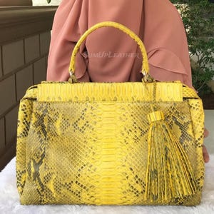 Könnte beinhalten: Eine gelbe Handtasche mit Schlangenmuster. Die Tasche hat einen oberen Griff, einen Klappenverschluss und eine dekorative Quaste. Die Tasche besteht aus einem strukturierten Material und hat eine rechteckige Form. Der Text "SUMUPLEATHER" ist sichtbar.