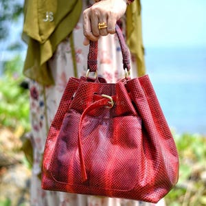 May include: A red snakeskin-effect handbag with a drawstring closure and a small external pocket. The bag has a structured shape with a rounded base and a gold-coloured buckle. The handles are also red and textured.