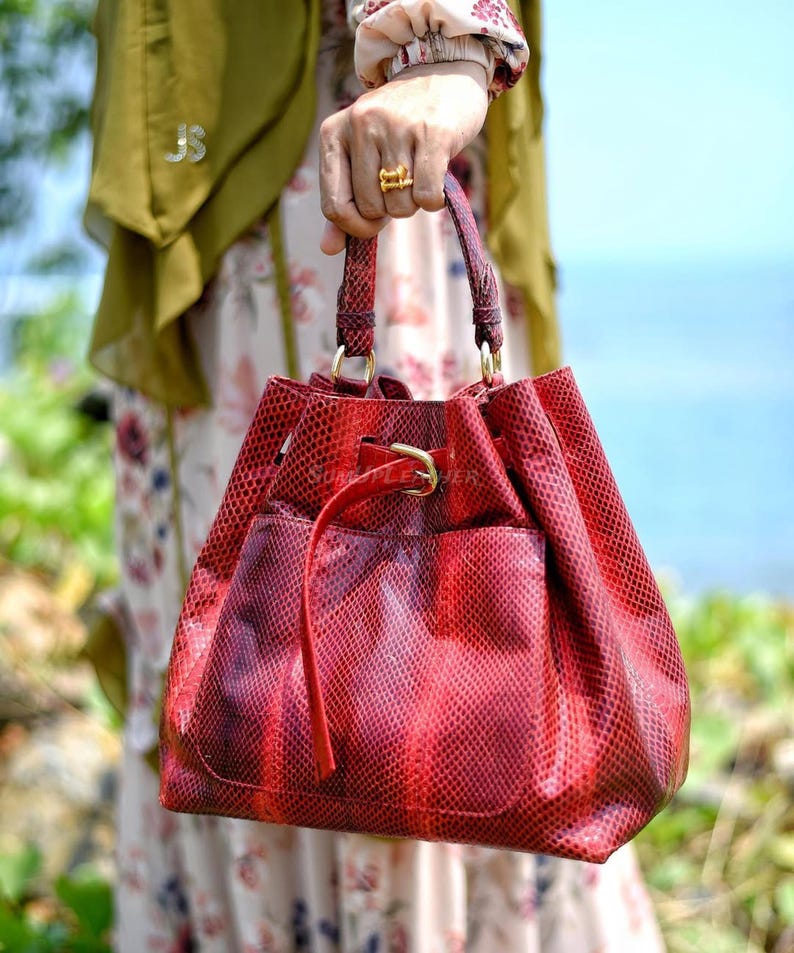 May include: A red snakeskin-effect handbag with a drawstring closure and a small external pocket. The bag has a structured shape with a rounded base and a gold-coloured buckle. The handles are also red and textured.