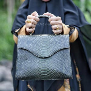 May include: A dark green snakeskin handbag with a curved flap closure. The bag has a top handle and a textured surface. The bag is held by a person wearing gold rings.