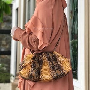 Könnte beinhalten: Eine Handtasche mit Schlangenmuster und Schulterriemen. Die Tasche hat eine Mischung aus Braun- und Gelbtönen mit einer strukturierten Oberfläche. Die Tasche wird von einer Person getragen, die ein pfirsichfarbenes Outfit trägt.