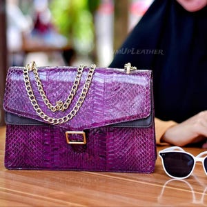 May include: A purple handbag with a snakeskin pattern, featuring a gold chain strap and clasp. The bag has a black top section and a gold rectangular buckle. The text "umUPLEATHER" is visible on the bag.