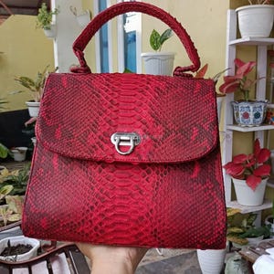 May include: A red snakeskin-effect handbag with a top handle and a silver clasp. The bag features a flap closure and a structured design. The overall aesthetic is bold and luxurious, suitable for various occasions.