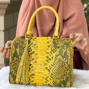 Könnte beinhalten: Gelb-schwarze Handtasche mit Schlangenmuster, gebogenem Griff und Quaste. Die rechteckige Tasche hat ein strukturiertes Design. Der Text "SUMUPLEATHER" ist sichtbar. Die Tasche wird von einer Person gehalten.